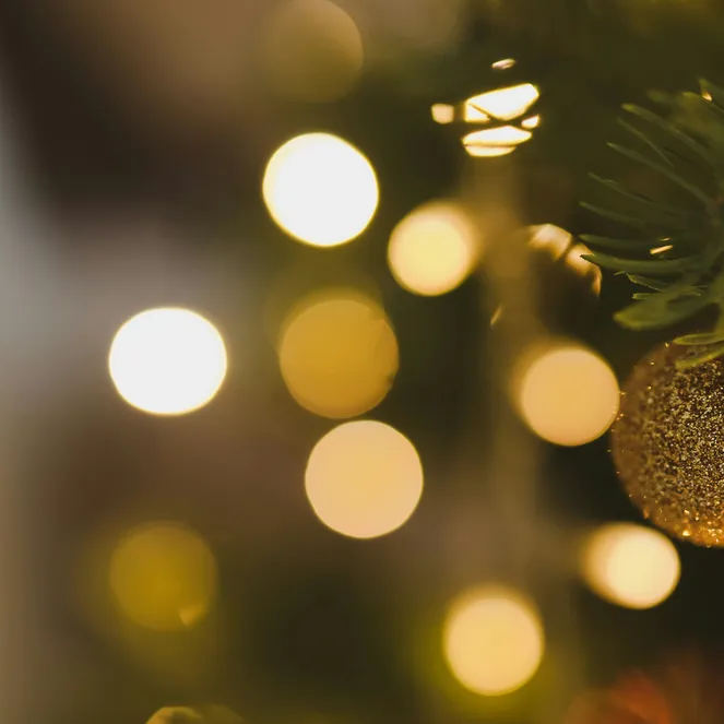 Close-up of a Christmas tree with a gold ornament. Blurred golden lights sparkle softly in the background.
