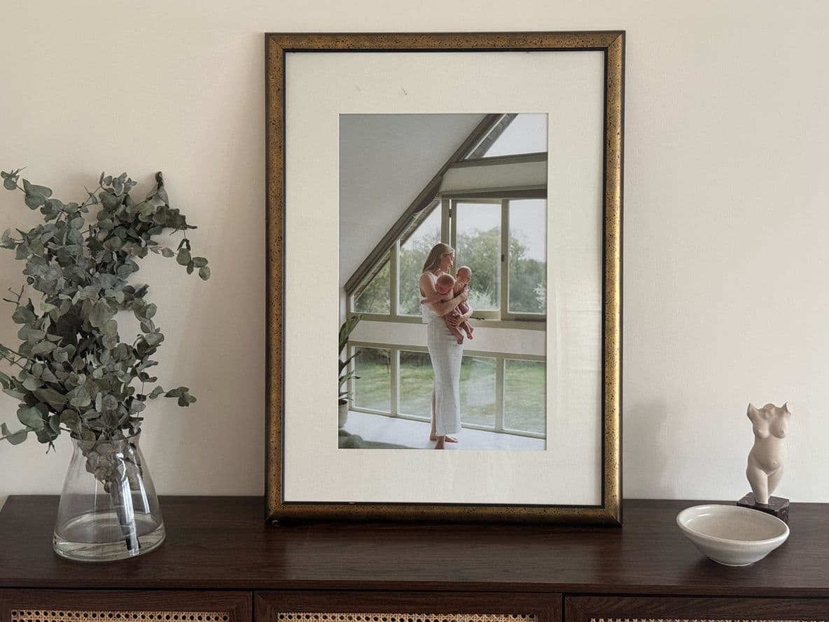 Framed photo on a wooden cabinet with a vase of greenery and a small bowl, against a plain wall.
