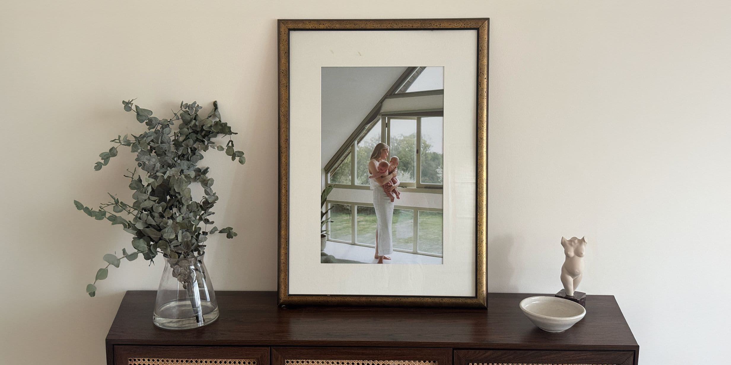 Framed photo on a wooden cabinet with a vase of greenery and a small bowl, against a plain wall.