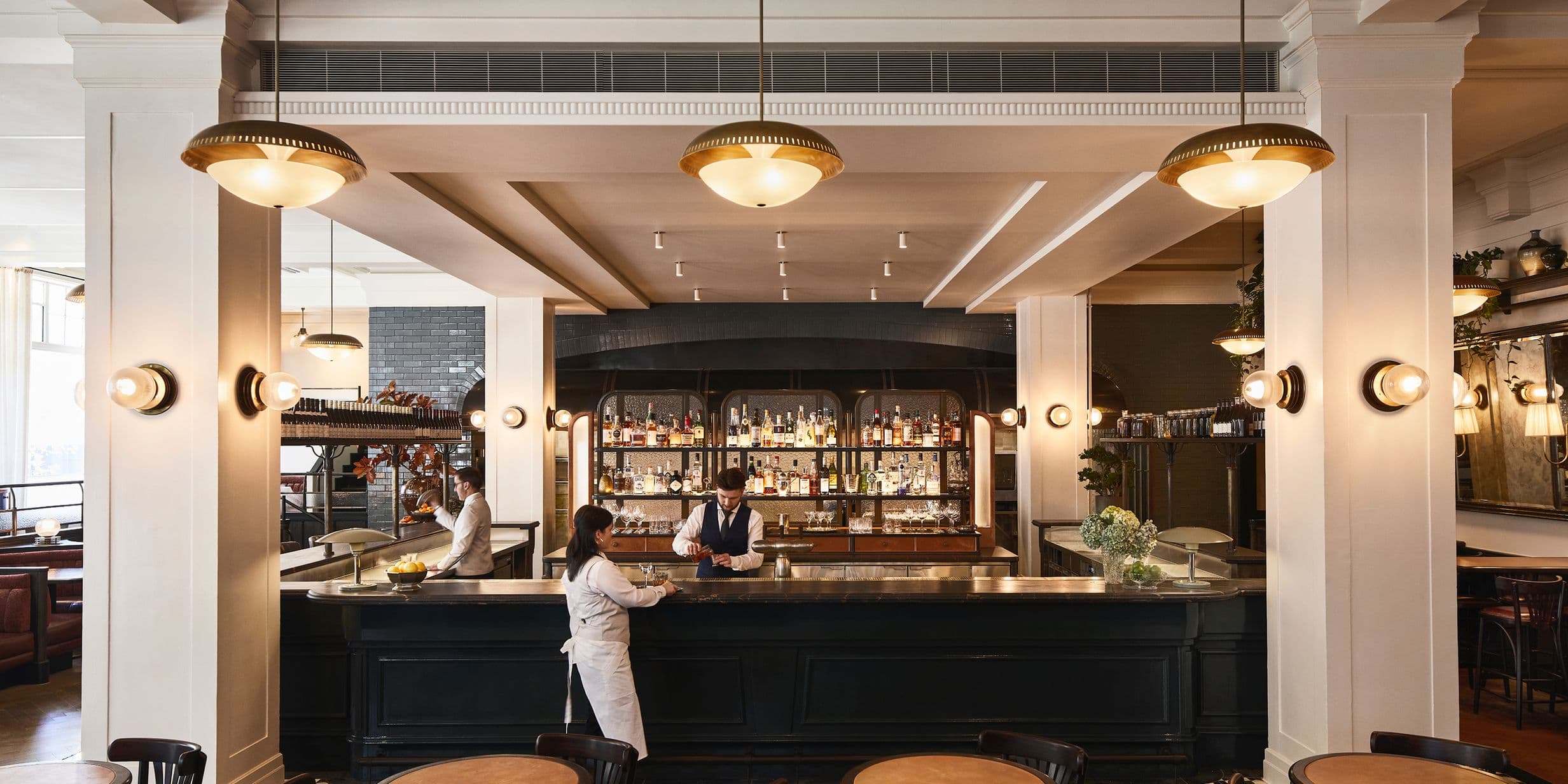 A spacious bar with elegant lighting, a bartender serves a customer. The background features a well-stocked bar with shelves of spirits and glasses.
