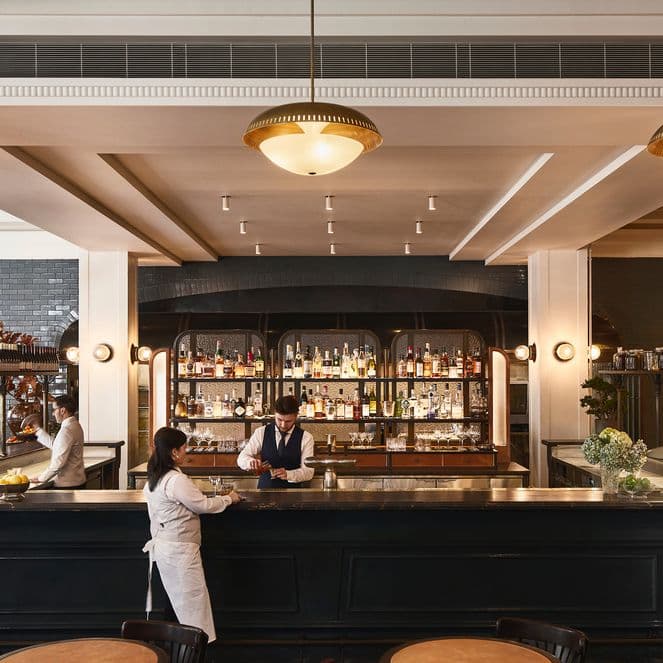 A spacious bar with elegant lighting, a bartender serves a customer. The background features a well-stocked bar with shelves of spirits and glasses.