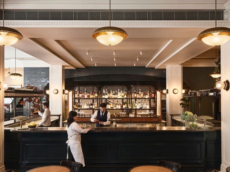 A spacious bar with elegant lighting, a bartender serves a customer. The background features a well-stocked bar with shelves of spirits and glasses.