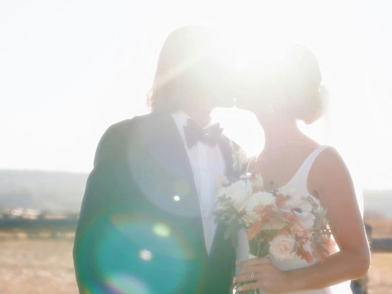 A couple in wedding attire kiss in bright sunlight, holding a bouquet, with lens flare and a blurred natural background.