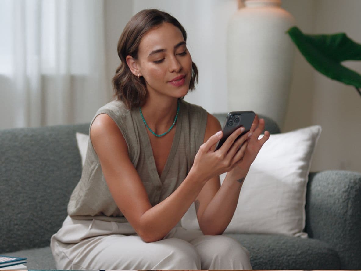 Woman sitting on a gray couch, looking at a smartphone, with a relaxed expression. Indoor setting with green plant in the background.