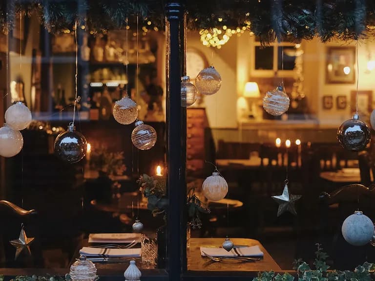 Cozy restaurant interior viewed through a window decorated with hanging ornaments and lights, tables set for dinner, and warm ambient lighting.