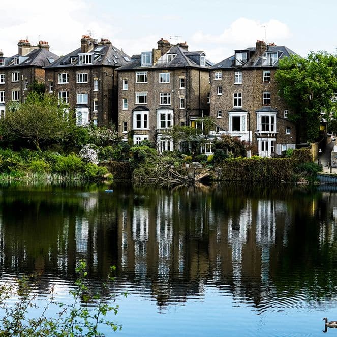 A row of brick houses reflected in a tranquil pond, surrounded by lush greenery and trees, with a single duck swimming in the foreground.