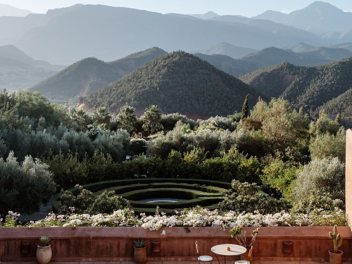 Terrace view of lush garden with circular design, surrounded by mountains under a clear sky. Rustic walls frame the scene.