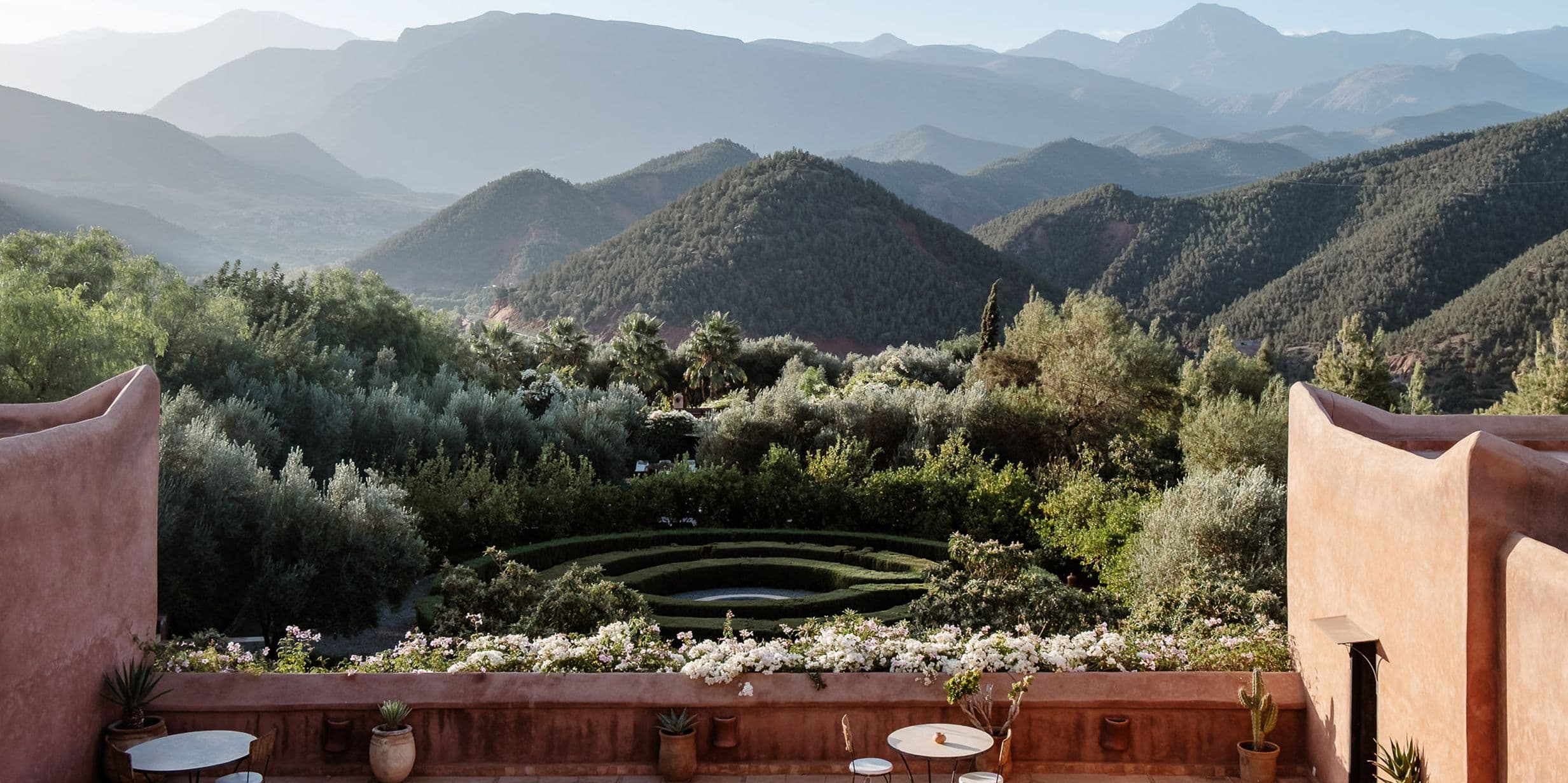 Terrace view of lush garden with circular design, surrounded by mountains under a clear sky. Rustic walls frame the scene.