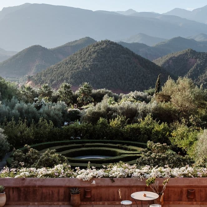 Terrace view of lush garden with circular design, surrounded by mountains under a clear sky. Rustic walls frame the scene.