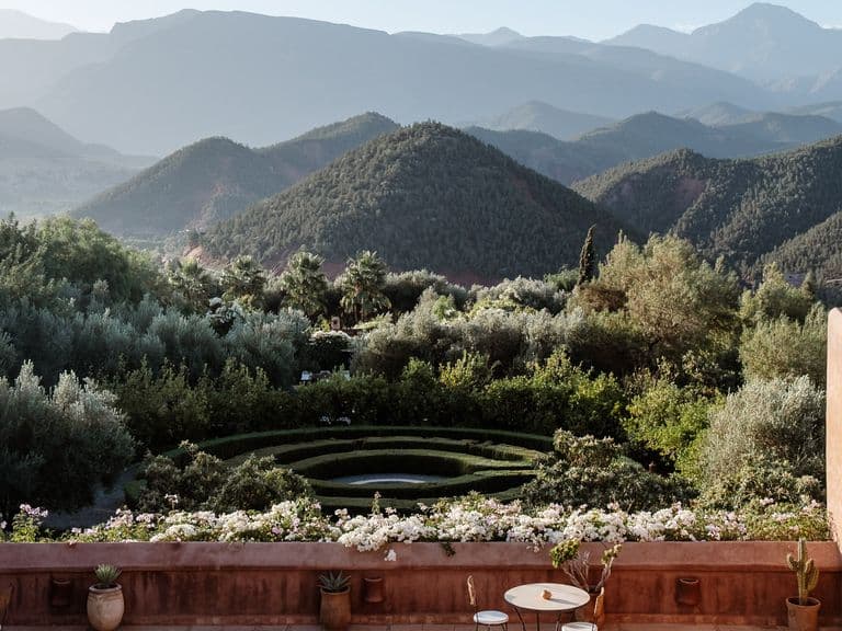 Terrace view of lush garden with circular design, surrounded by mountains under a clear sky. Rustic walls frame the scene.