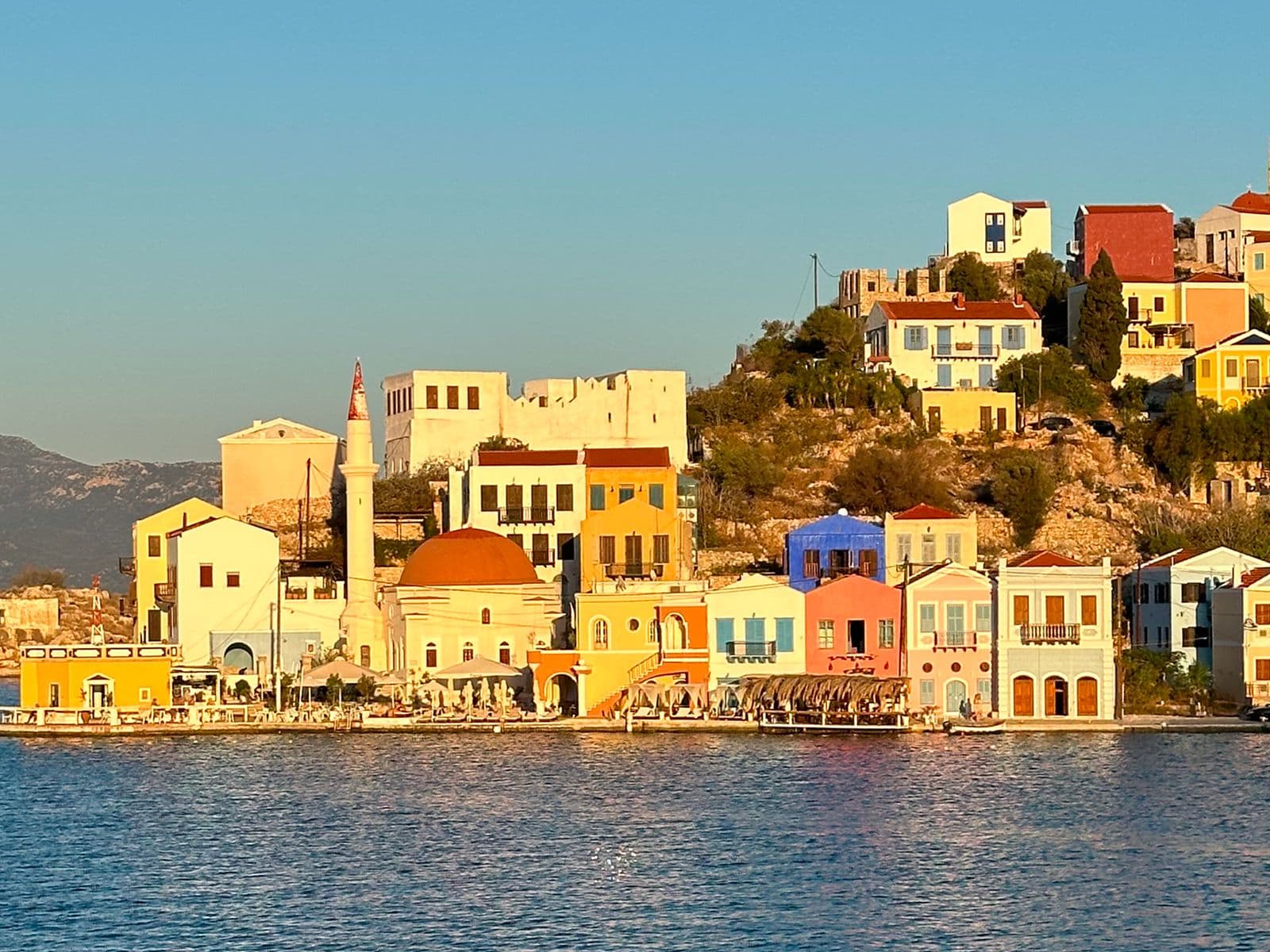 Colorful houses on a sunlit hillside by the sea, with a clear blue sky and distant mountains in the background.
