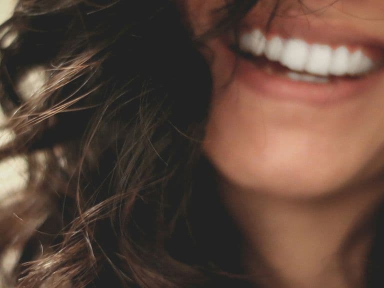 Close-up of a person smiling with white teeth, slightly curled dark hair, and blurred background.