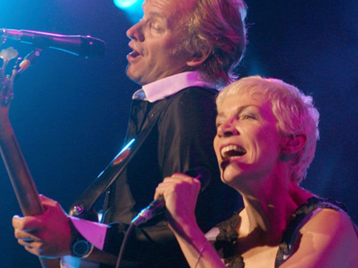 Two performers singing passionately on stage, one playing guitar. Blue stage lights illuminate the scene.