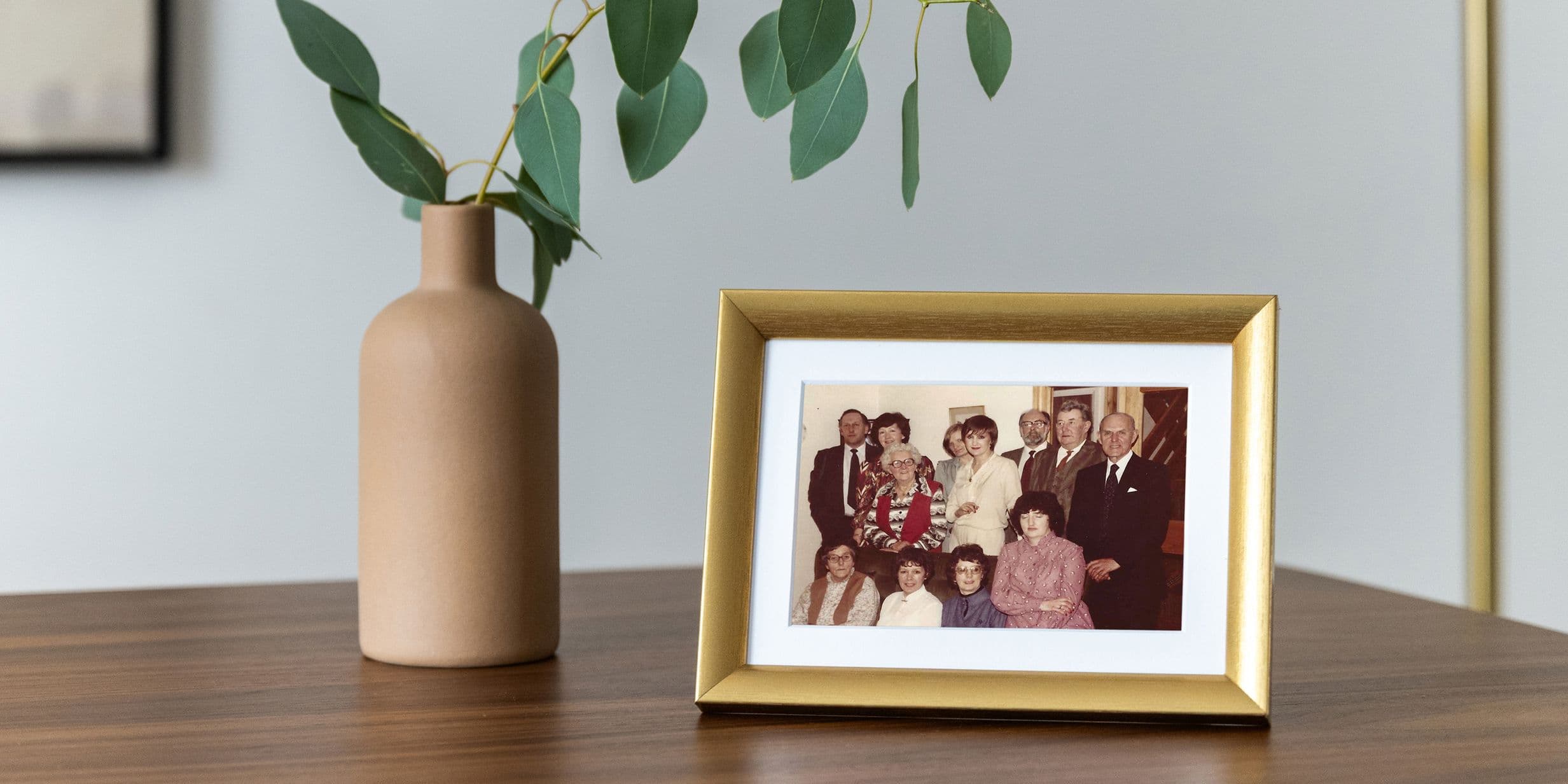 A framed family photo on a wooden table next to a beige vase with green leaves.