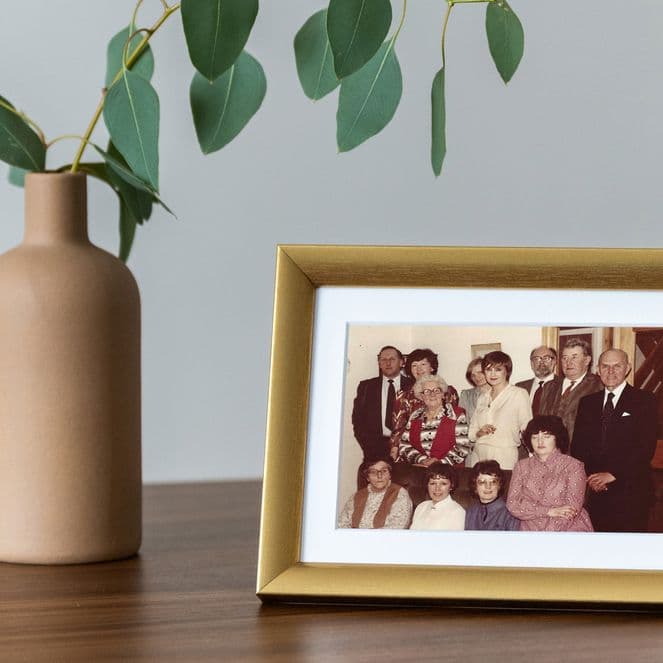 A framed family photo on a wooden table next to a beige vase with green leaves.