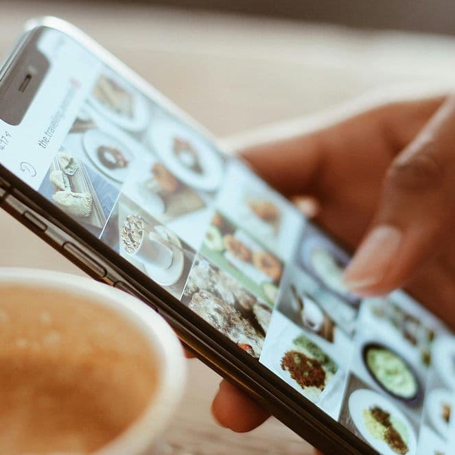 A hand holding a smartphone, browsing through food photos, with a cup of coffee on a wooden table in the background.