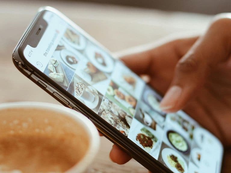 A hand holding a smartphone, browsing through food photos, with a cup of coffee on a wooden table in the background.