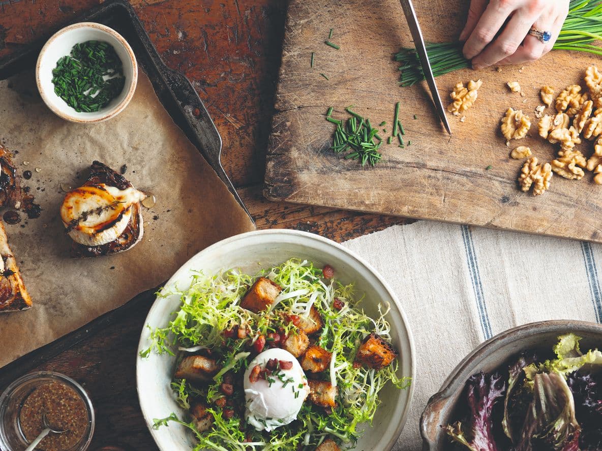 A rustic dining table with a salad, bruschetta, nuts, and chopped herbs. A hand is chopping chives on a wooden board.