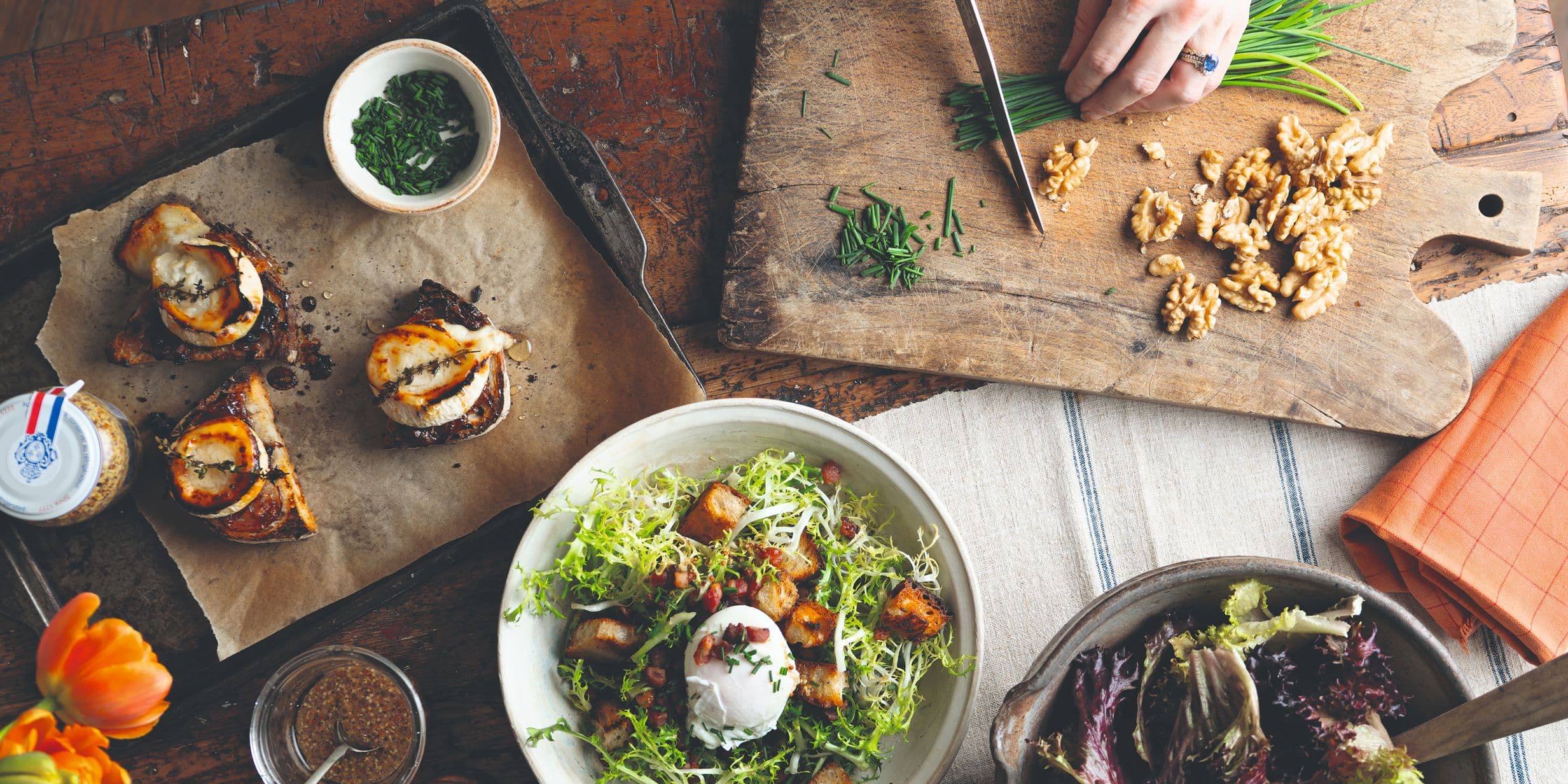A rustic dining table with a salad, bruschetta, nuts, and chopped herbs. A hand is chopping chives on a wooden board.