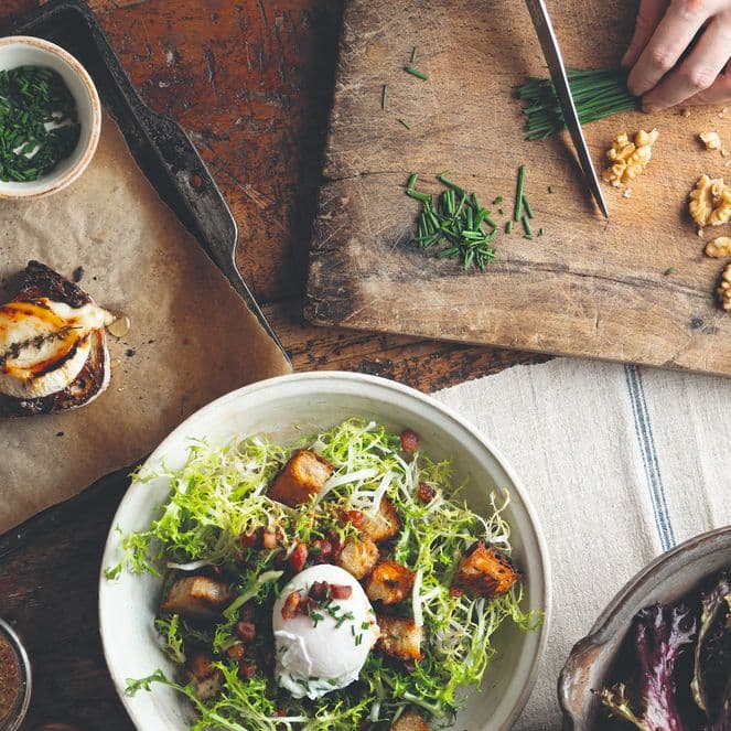 A rustic dining table with a salad, bruschetta, nuts, and chopped herbs. A hand is chopping chives on a wooden board.