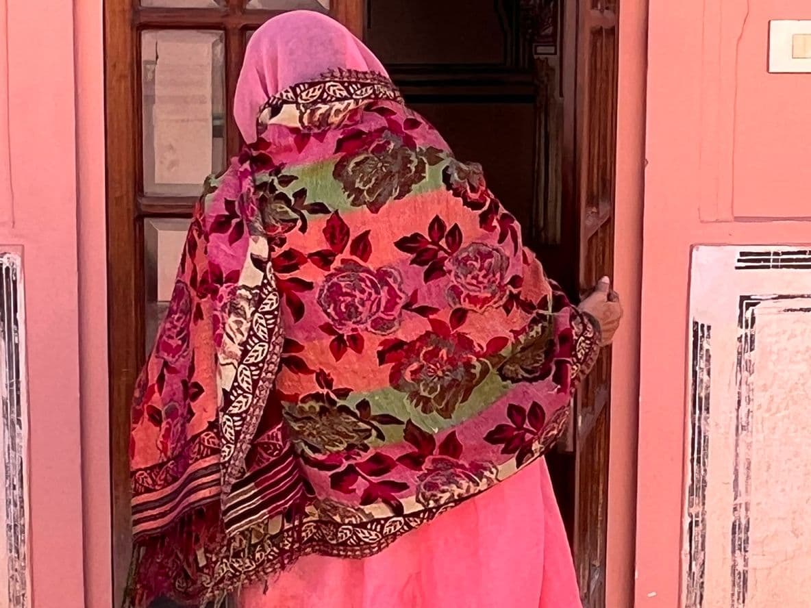 A person in a colorful floral shawl and pink attire enters a building with a pink wall and white decorative accents.