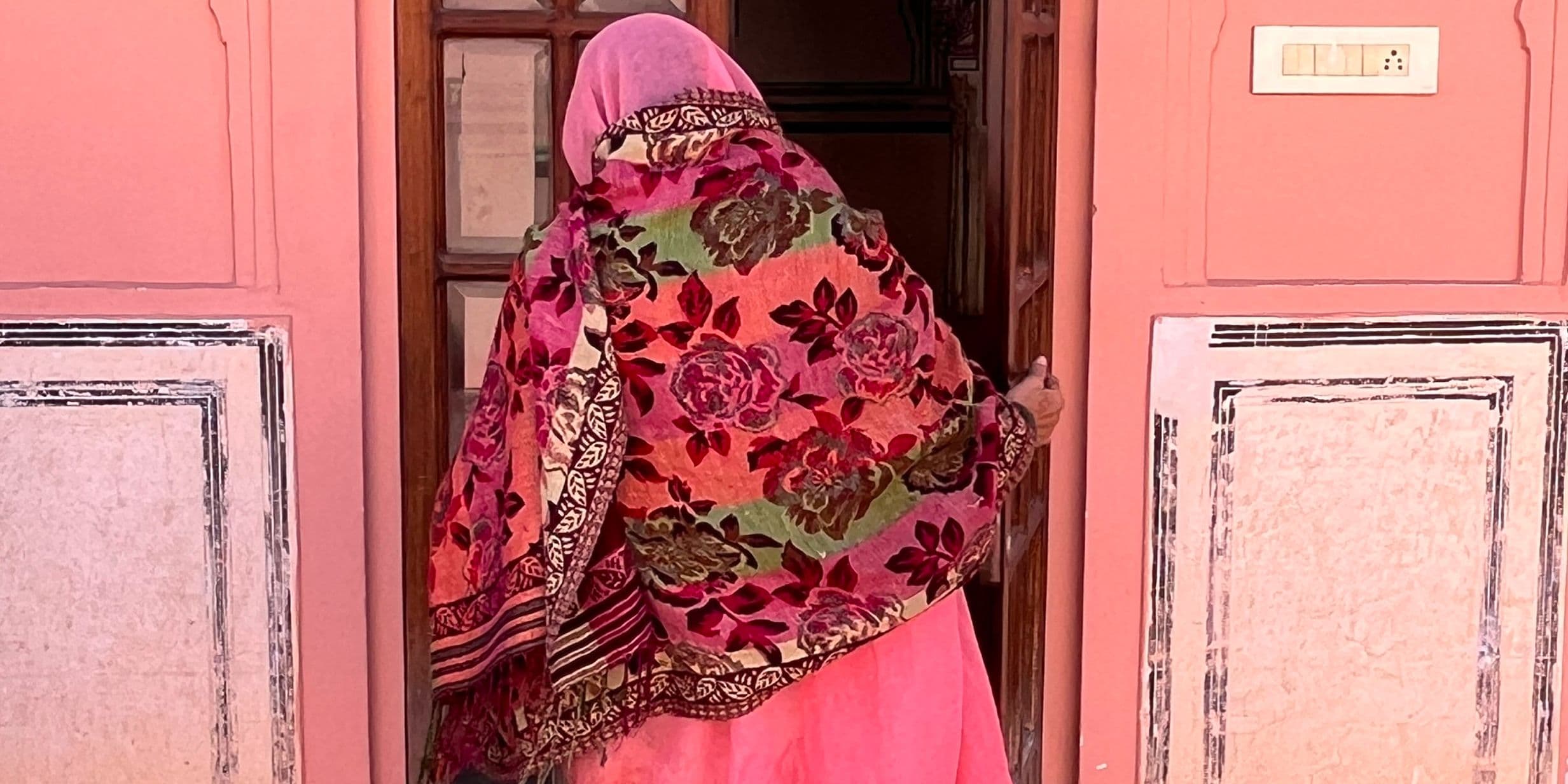 A person in a colorful floral shawl and pink attire enters a building with a pink wall and white decorative accents.
