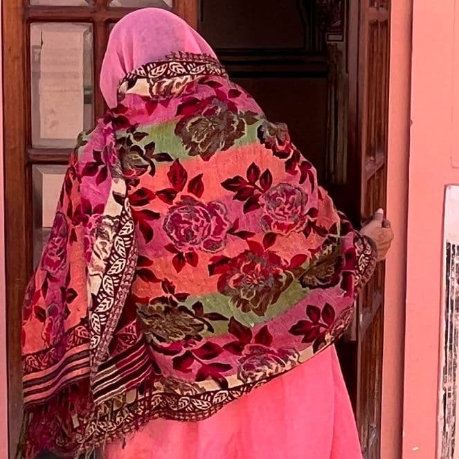 A person in a colorful floral shawl and pink attire enters a building with a pink wall and white decorative accents.