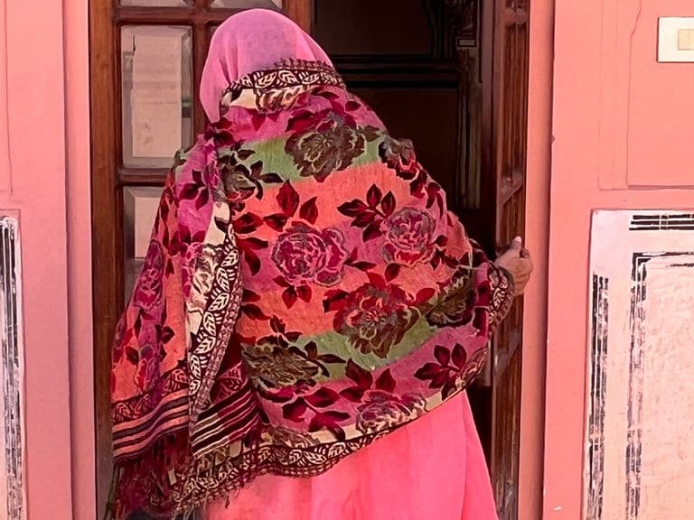 A person in a colorful floral shawl and pink attire enters a building with a pink wall and white decorative accents.