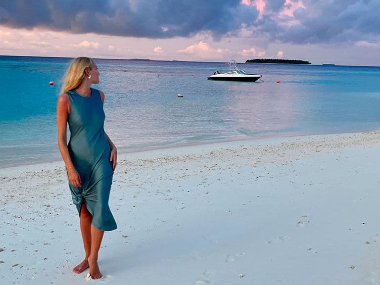 A woman in a blue dress stands on a beach at sunset, with turquoise waters and a boat in the distance under a cloudy sky.
