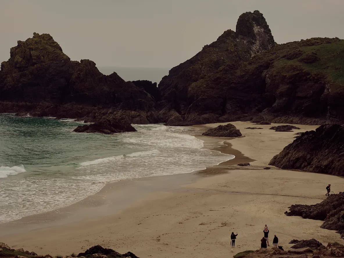 A picturesque beach with large rocky formations, gentle waves, sandy shore, and a group of people exploring under a cloudy sky.