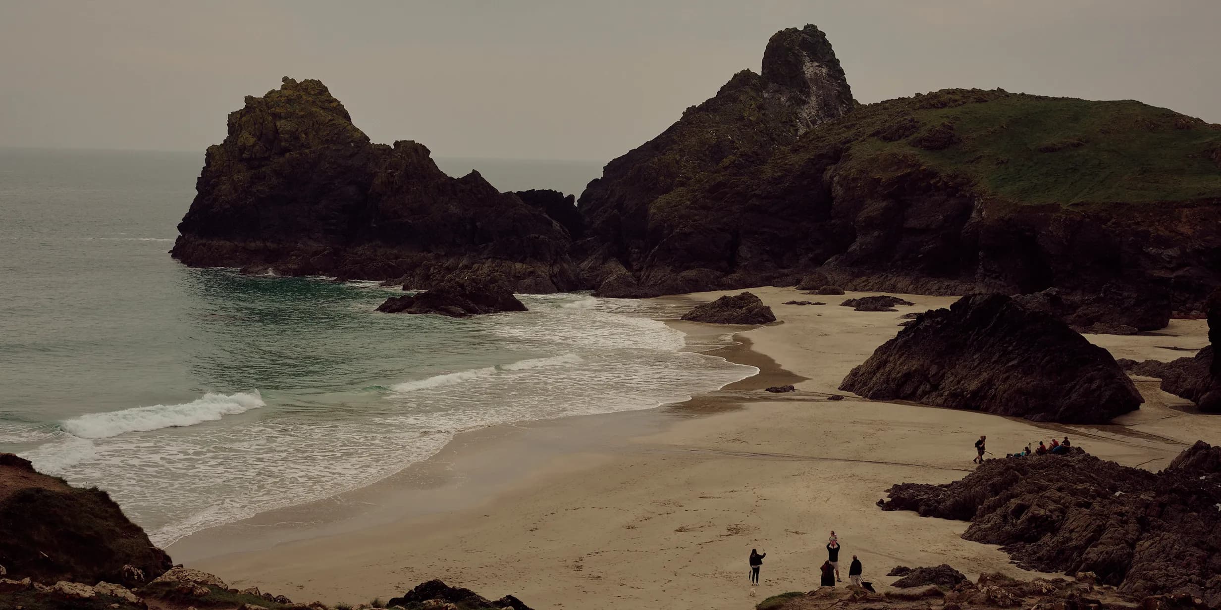 A picturesque beach with large rocky formations, gentle waves, sandy shore, and a group of people exploring under a cloudy sky.