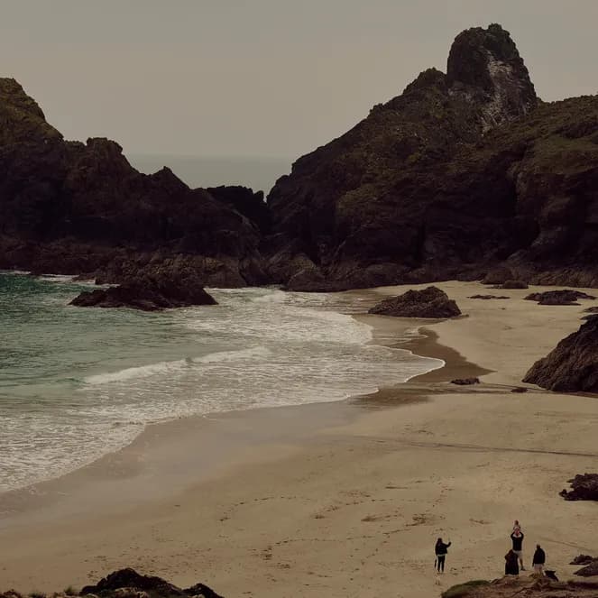 A picturesque beach with large rocky formations, gentle waves, sandy shore, and a group of people exploring under a cloudy sky.