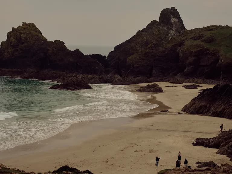 A picturesque beach with large rocky formations, gentle waves, sandy shore, and a group of people exploring under a cloudy sky.