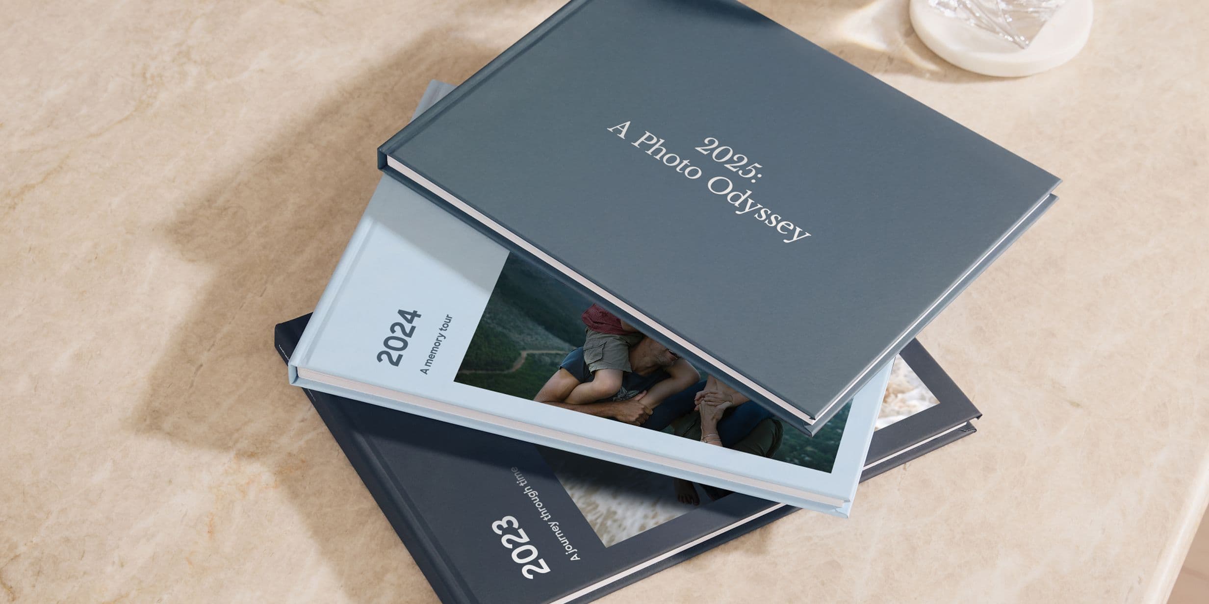 Three photo books titled "2023," "2024," and "2025: A Photo Odyssey" are stacked on a marble surface.