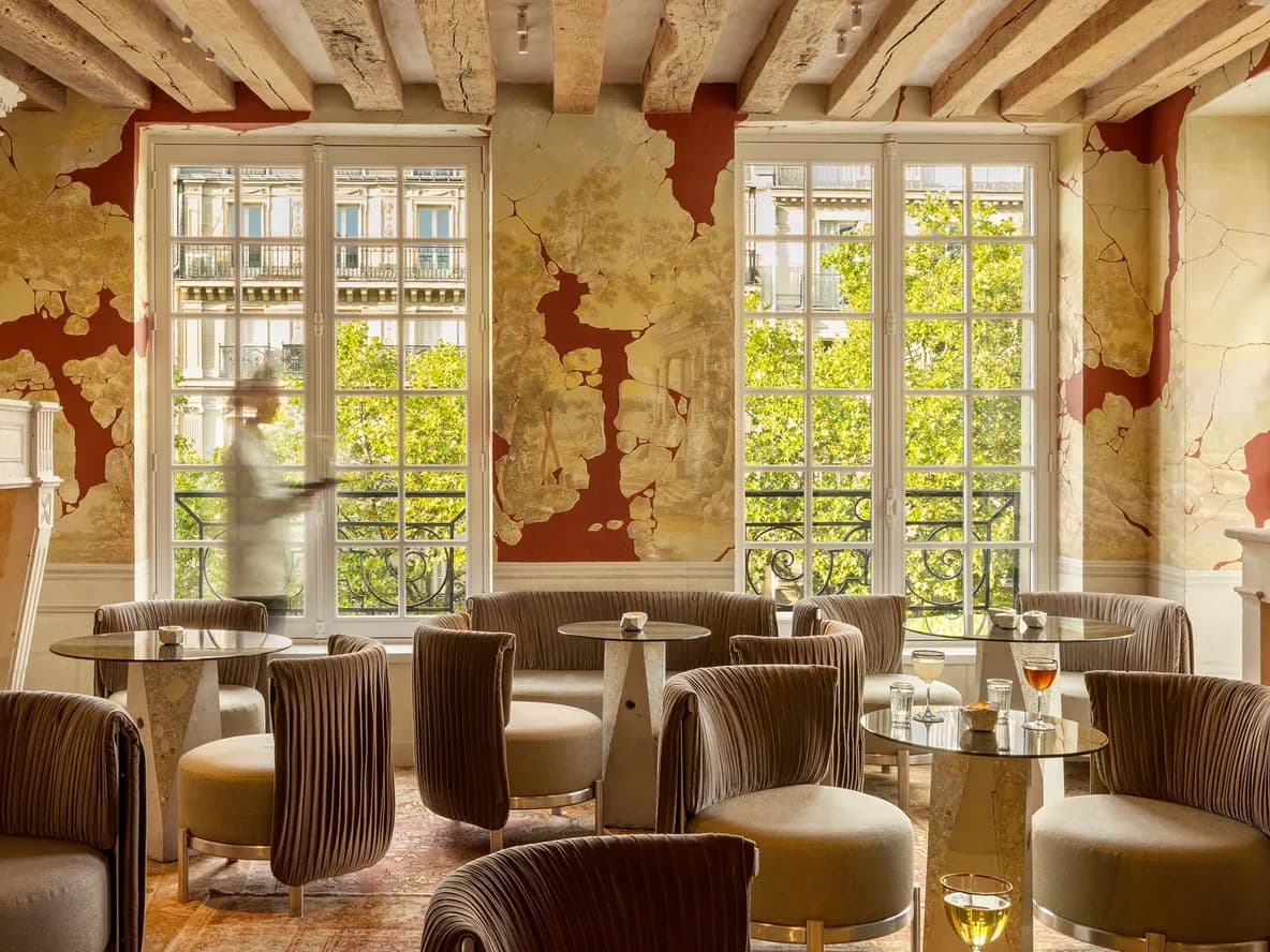 Elegant lounge with plush chairs, round tables, and drinks. Large windows reveal trees outside. Rustic decor with exposed wooden beams and ornate fireplace.