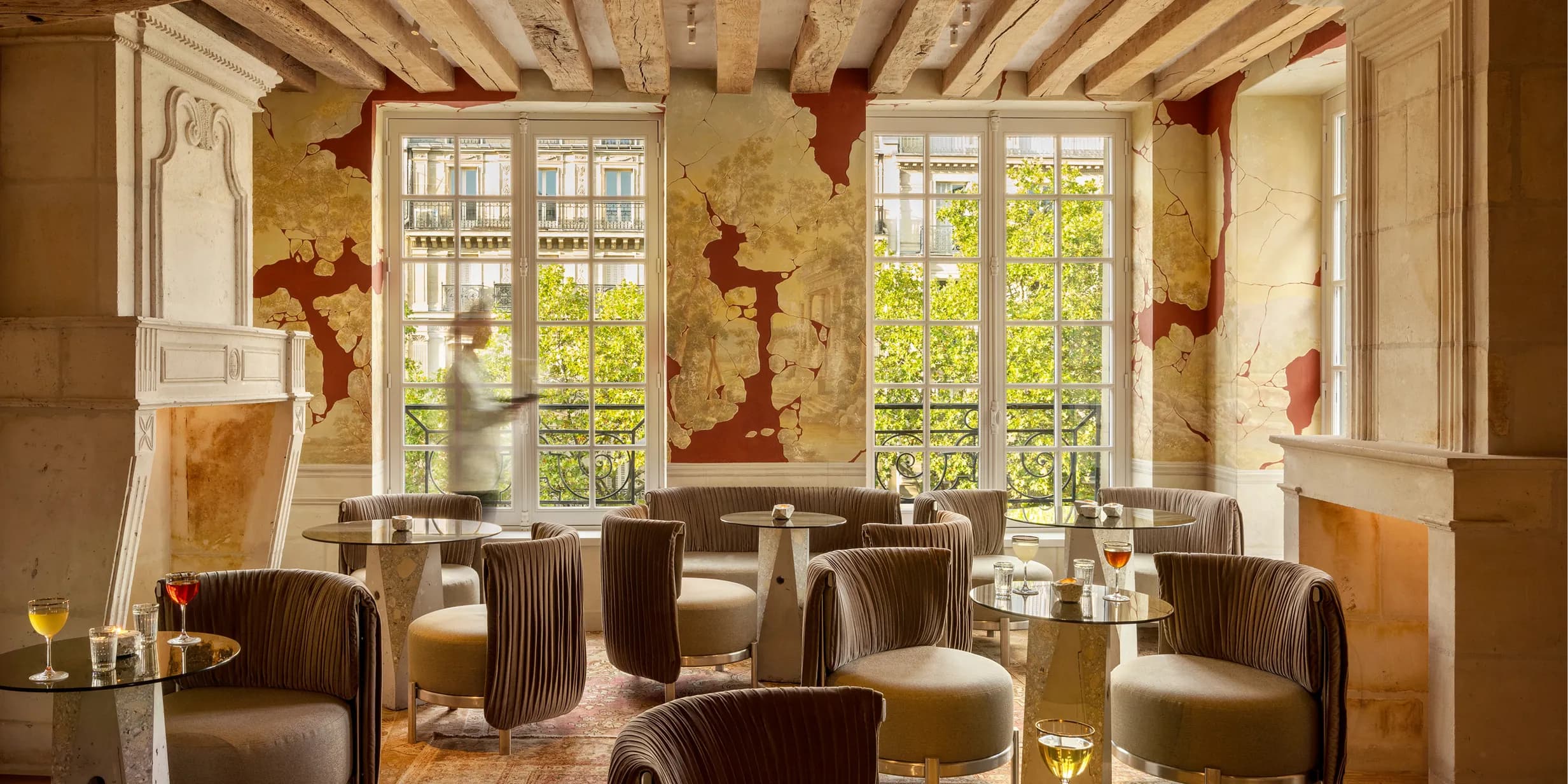 Elegant lounge with plush chairs, round tables, and drinks. Large windows reveal trees outside. Rustic decor with exposed wooden beams and ornate fireplace.