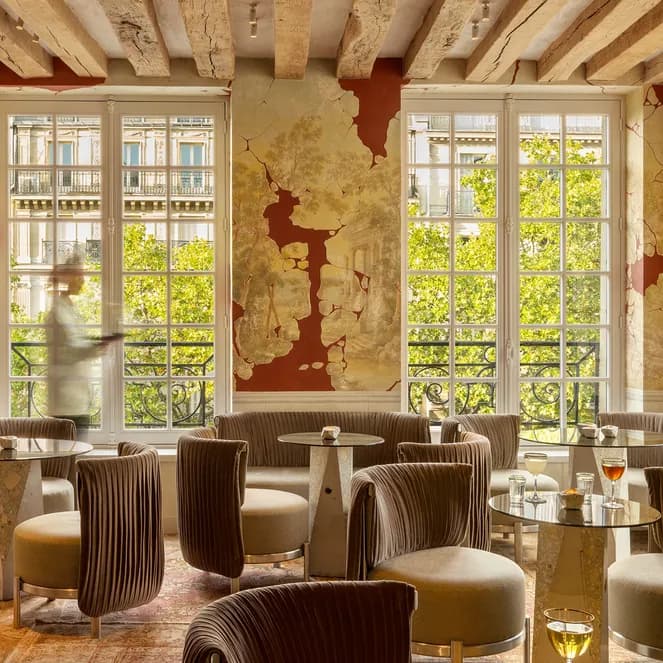 Elegant lounge with plush chairs, round tables, and drinks. Large windows reveal trees outside. Rustic decor with exposed wooden beams and ornate fireplace.