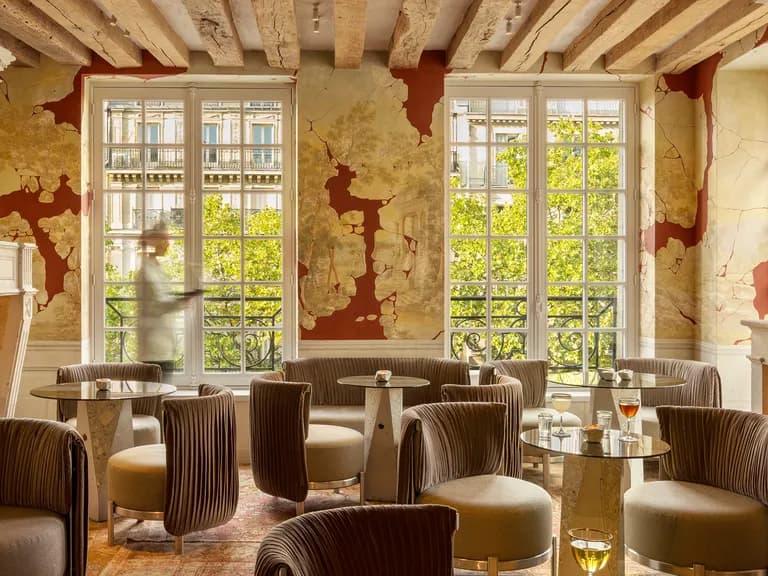 Elegant lounge with plush chairs, round tables, and drinks. Large windows reveal trees outside. Rustic decor with exposed wooden beams and ornate fireplace.