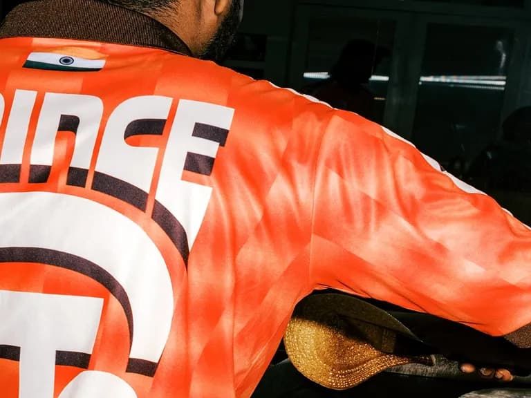 Man wearing an orange shirt with "PRINCE" printed on it, holding a shoe, reflected in a mirror.