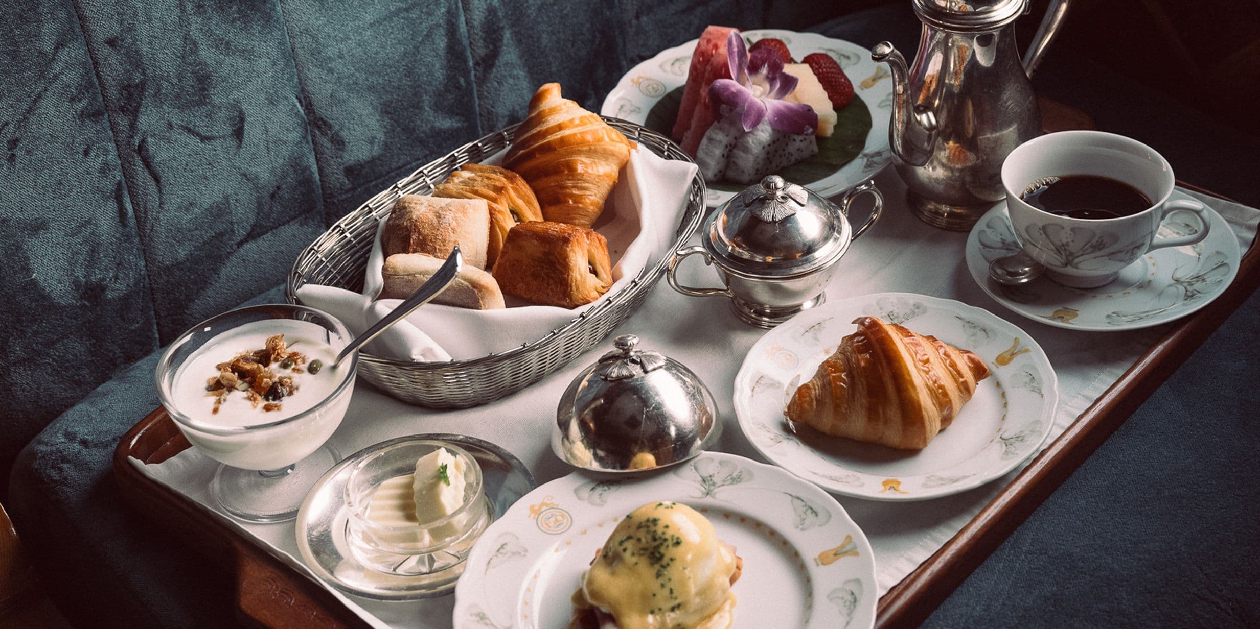 A breakfast tray with croissants, rolls, yogurt, butter, a poached egg dish, fruit salad, and a cup of coffee, set on a plush surface.