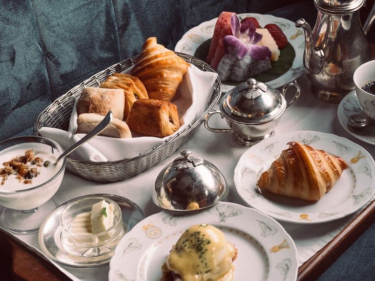 A breakfast tray with croissants, rolls, yogurt, butter, a poached egg dish, fruit salad, and a cup of coffee, set on a plush surface.