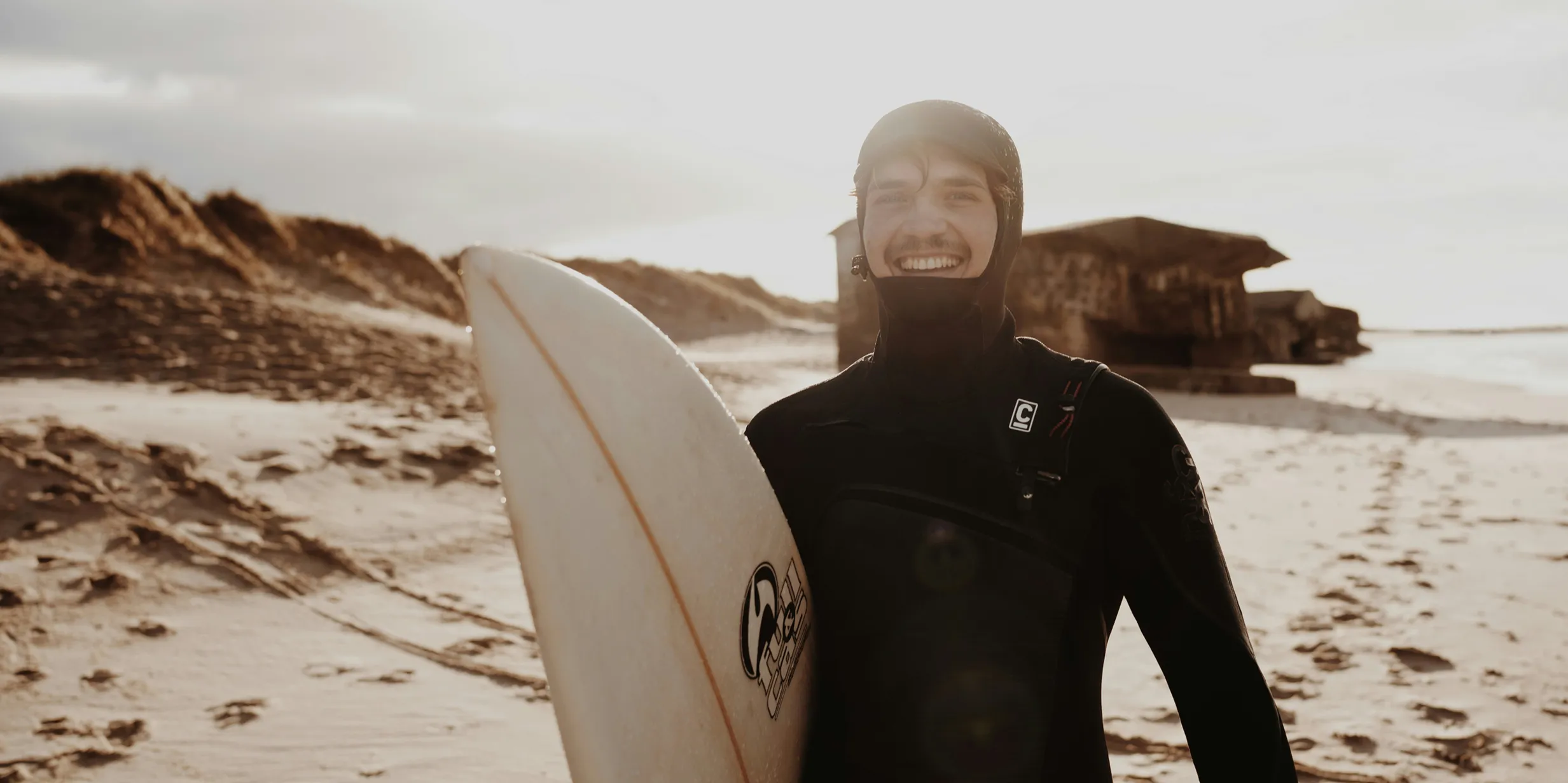 Surfer in a wetsuit smiling and holding a surfboard on a sandy beach with dunes and a wooden hut in the background.