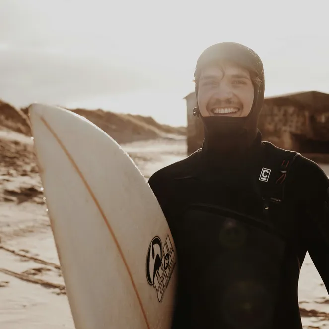 Surfer in a wetsuit smiling and holding a surfboard on a sandy beach with dunes and a wooden hut in the background.