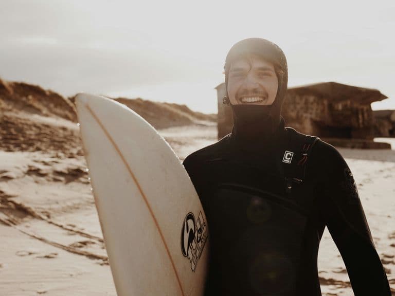 Surfer in a wetsuit smiling and holding a surfboard on a sandy beach with dunes and a wooden hut in the background.