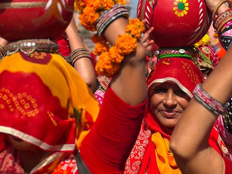 Women in vibrant traditional attire carry decorated pots on their heads during a cultural procession, adorned with floral garlands and bangles.