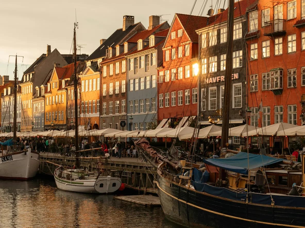 Colorful buildings and moored boats along a canal in Nyhavn, Copenhagen, under warm, golden evening light, with lively outdoor cafes.