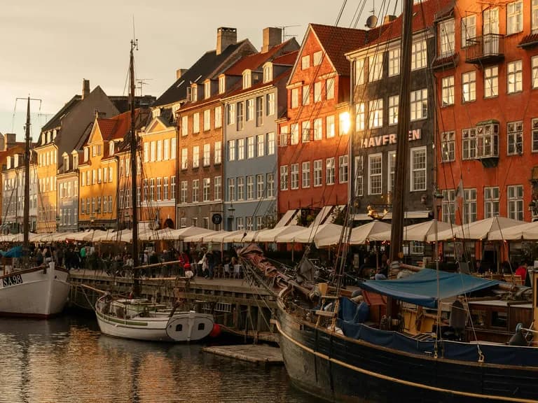 Colorful buildings and moored boats along a canal in Nyhavn, Copenhagen, under warm, golden evening light, with lively outdoor cafes.
