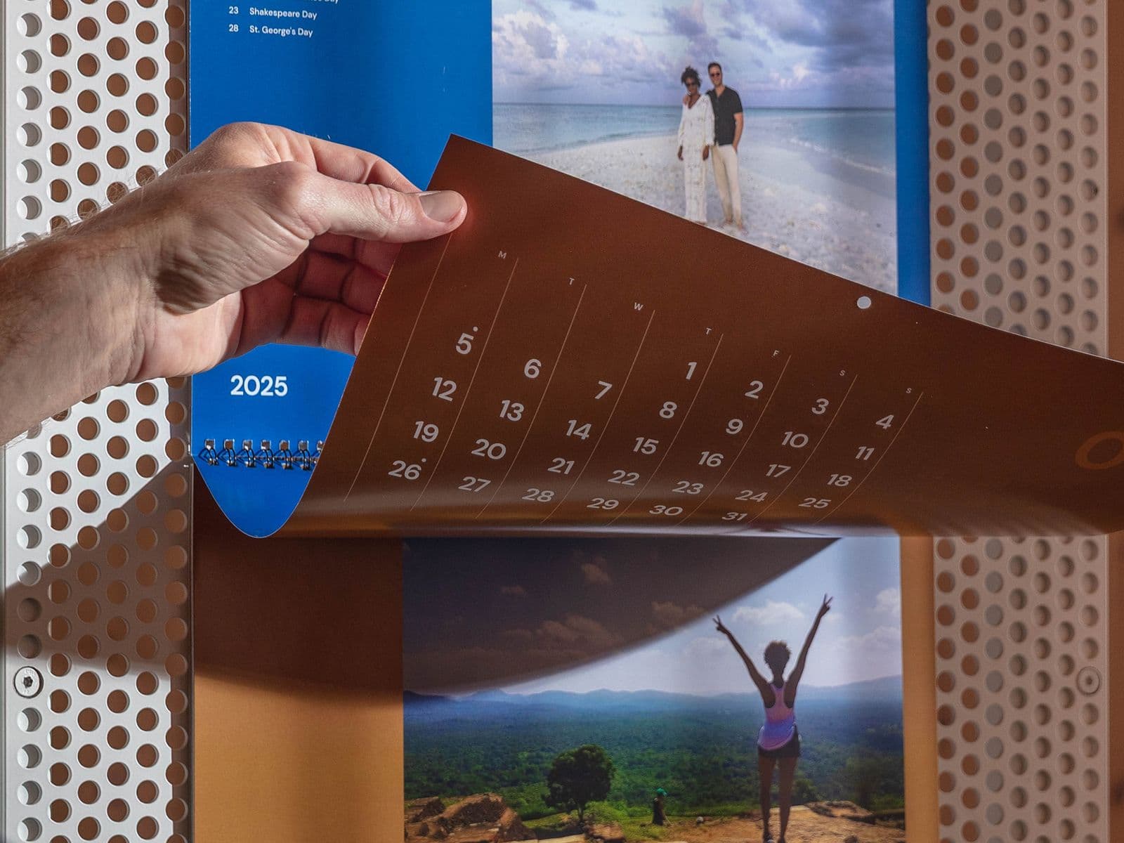 Person flipping calendar page to May 2025, featuring beach and mountain images, on a perforated wall background.