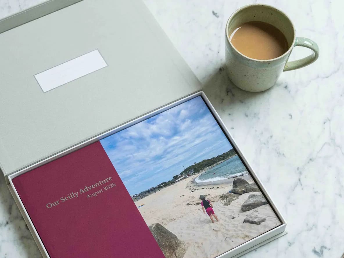 A photo book labeled "Our Scilly Adventure August 2026" lies open in a box on a marble table next to a mug of tea.