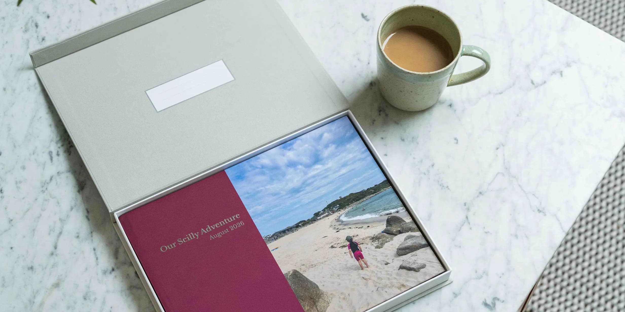 A photo book labeled "Our Scilly Adventure August 2026" lies open in a box on a marble table next to a mug of tea.